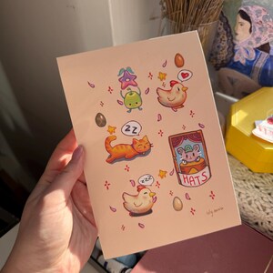 May include: A light pink art print featuring whimsical illustrations of a sleeping orange cat, a sleeping chicken, and other cute characters. The print includes the text "HATS" and the artist's signature.