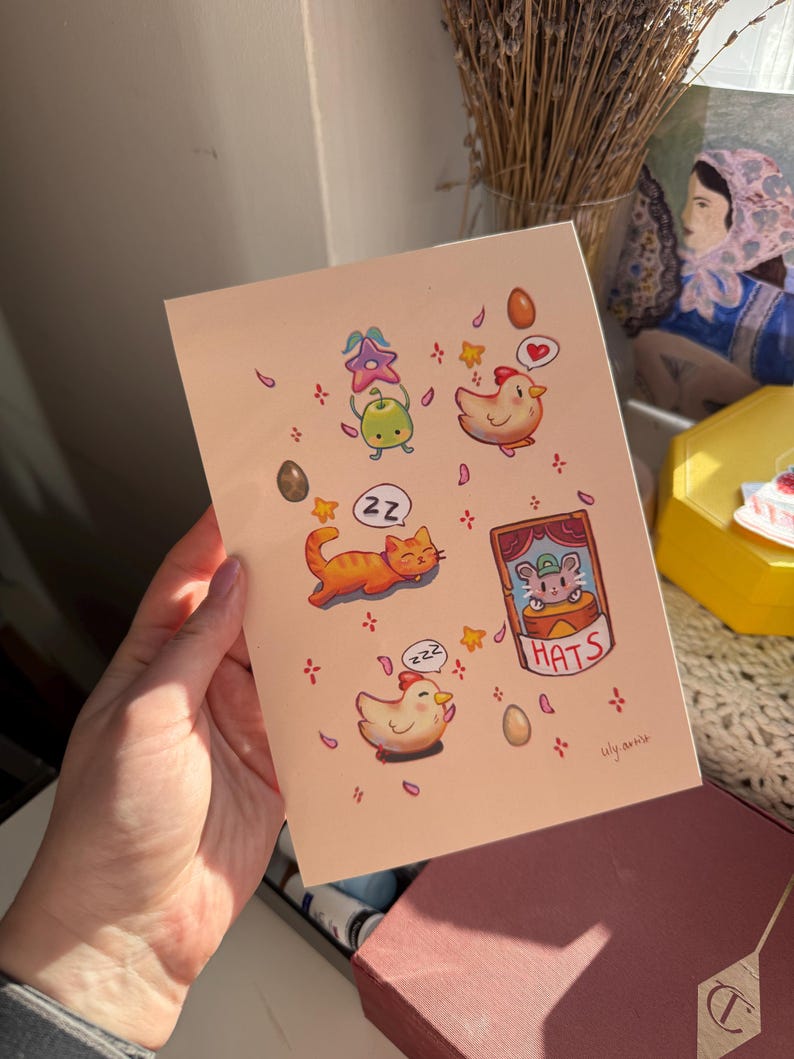 May include: A light pink art print featuring whimsical illustrations of a sleeping orange cat, a sleeping chicken, and other cute characters. The print includes the text "HATS" and the artist's signature.
