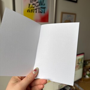 Op de afbeelding: Open witte blanco wenskaart met een gestippelde papiertextuur. De kaart wordt opengehouden en toont de binnenpagina's. Op de achtergrond staat een ingelijste poster met de tekst "SUPPORT YOUR LOCAL ARTIST".