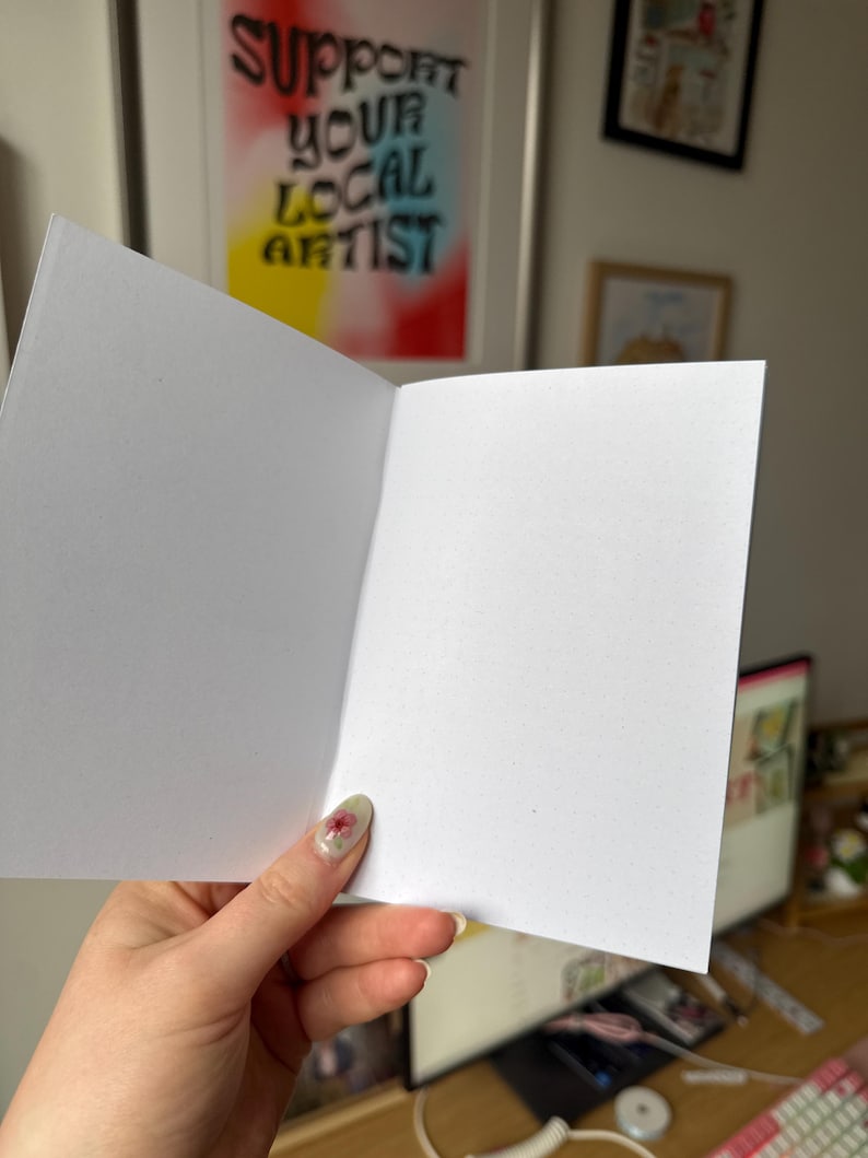 Op de afbeelding: Open witte blanco wenskaart met een gestippelde papiertextuur. De kaart wordt opengehouden en toont de binnenpagina's. Op de achtergrond staat een ingelijste poster met de tekst "SUPPORT YOUR LOCAL ARTIST".