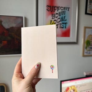 Op de afbeelding: Een lichtroze notitieboekje met een kleurrijke lolly-afbeelding in de hoek. Het notitieboekje wordt vastgehouden voor een muur met ingelijste kunst, waaronder een stuk met de tekst "SUPPORT YOUR LOCAL ARTIST".