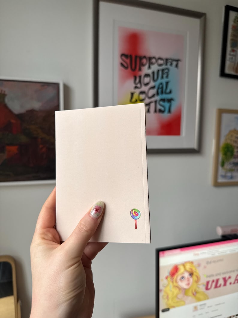 Op de afbeelding: Een lichtroze notitieboekje met een kleurrijke lolly-afbeelding in de hoek. Het notitieboekje wordt vastgehouden voor een muur met ingelijste kunst, waaronder een stuk met de tekst "SUPPORT YOUR LOCAL ARTIST".