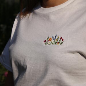 Puede incluir: Camiseta blanca con un dise&ntilde;o floral bordado en el pecho. El bordado presenta una variedad de flores coloridas, incluyendo rojas, amarillas, azules y naranjas, con tallos y hojas verdes.