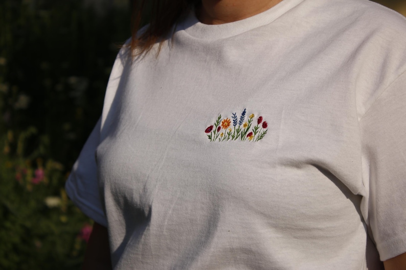 Puede incluir: Camiseta blanca con un dise&ntilde;o floral bordado en el pecho. El bordado presenta una variedad de flores coloridas, incluyendo rojas, amarillas, azules y naranjas, con tallos y hojas verdes.