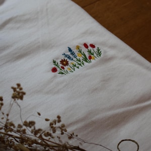 Puede incluir: Camiseta blanca con un dise&ntilde;o floral bordado. El bordado presenta una variedad de flores coloridas, incluyendo rojas, amarillas y azules, dispuestas en forma rectangular. La camiseta est&aacute; hecha de una tela suave y clara.