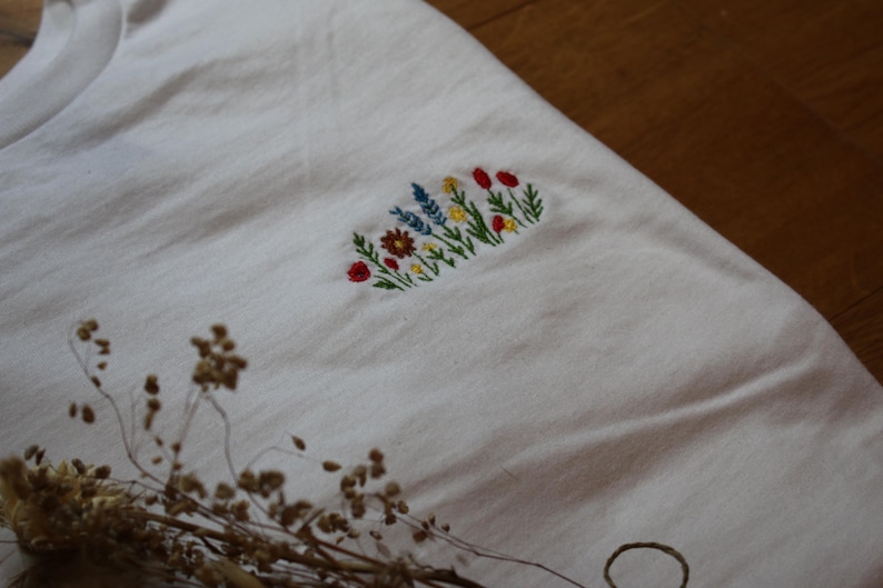 Puede incluir: Camiseta blanca con un dise&ntilde;o floral bordado. El bordado presenta una variedad de flores coloridas, incluyendo rojas, amarillas y azules, dispuestas en forma rectangular. La camiseta est&aacute; hecha de una tela suave y clara.
