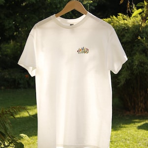 Puede incluir: Camiseta blanca con un peque&ntilde;o bordado floral colorido en el pecho. La camiseta est&aacute; colgada de una percha de madera sobre un fondo de follaje verde y hierba. La camiseta tiene mangas cortas y cuello redondo.