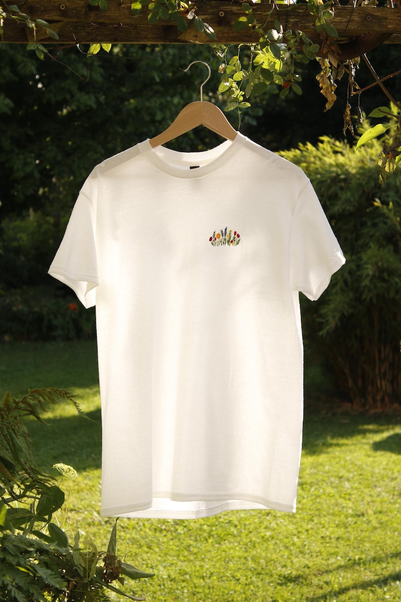 Puede incluir: Camiseta blanca con un peque&ntilde;o bordado floral colorido en el pecho. La camiseta est&aacute; colgada de una percha de madera sobre un fondo de follaje verde y hierba. La camiseta tiene mangas cortas y cuello redondo.