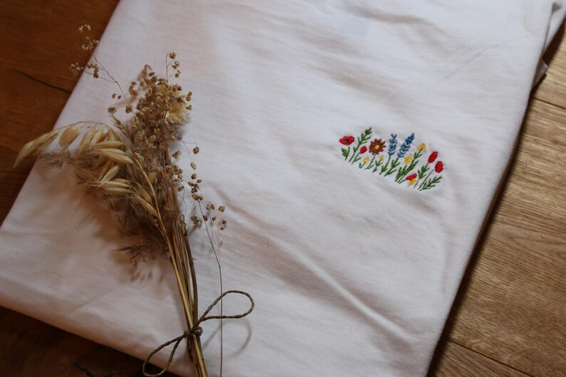 Puede incluir: Tela blanca con un dise&ntilde;o floral bordado en rojo, amarillo, azul y verde. Un manojo de trigo y hierbas secas, atado con hilo, descansa sobre la tela, sobre una superficie de madera.