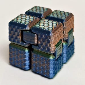 Può includere: Un cubo fidget multicolore stampato in 3D. Il cubo presenta un motivo a nido d'ape sulle sue superfici, con una sfumatura di colore che va dal blu al bronzo. Il cubo è progettato con segmenti ad incastro, che consentono movimenti e manipolazioni flessibili.