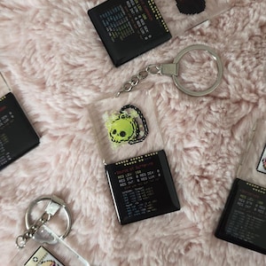 Könnte beinhalten: Schlüsselanhänger mit pixeligen Grafiken und Text. Die Schlüsselanhänger zeigen verschiedene Designs, darunter einen Schädel, eine rote Form und eine Figur. Jeder Schlüsselanhänger hat einen silbernen Schlüsselring. Die Schlüsselanhänger liegen auf einer rosa, flauschigen Oberfläche.