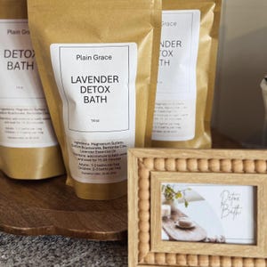 May include: Kraft paper bags of Lavender Detox Bath with white labels. The labels read "Plain Grace" and "Lavender Detox Bath." A small wooden frame with a photo of a bath scene is in the foreground. The bags are 14 oz.