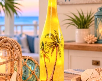 Op de afbeelding: Een hoge, cilindrische glazen fles die van binnenuit wordt verlicht met een warme gele gloed. Het buitenoppervlak is voorzien van een geschilderde illustratie van twee palmbomen op een zandstrand. De fles heeft een smalle hals en een ronde voet.