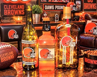 May include: Three glass bottles decorated with Cleveland Browns football helmet logos and team colors. The bottles are illuminated with internal string lights. The display includes a football, a coaster, and a bowl, all featuring the team logo.