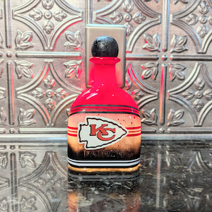 May include: A red glass bottle with a black top and a white, red, and black Kansas City Chiefs logo. The bottle has a black and white stripe pattern around the bottom. The text "PATRIOTS" is visible on the bottle.