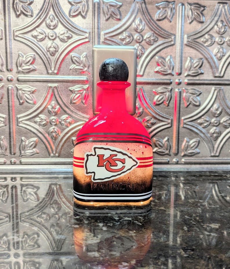 May include: A red glass bottle with a black top and a white, red, and black Kansas City Chiefs logo. The bottle has a black and white stripe pattern around the bottom. The text "PATRIOTS" is visible on the bottle.