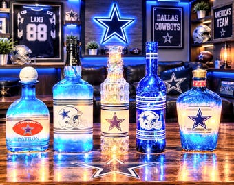 May include: Five decorative glass bottles illuminated with internal lights, each featuring Dallas Cowboys branding. The collection includes various shapes and sizes, showcasing team logos, helmets, and stars in blue and white, arranged on a wooden surface.