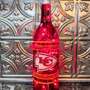 May include: A red glass wine bottle with a white and red Kansas City Chiefs logo. The bottle has orange stripes around it and is lit from inside with white lights.