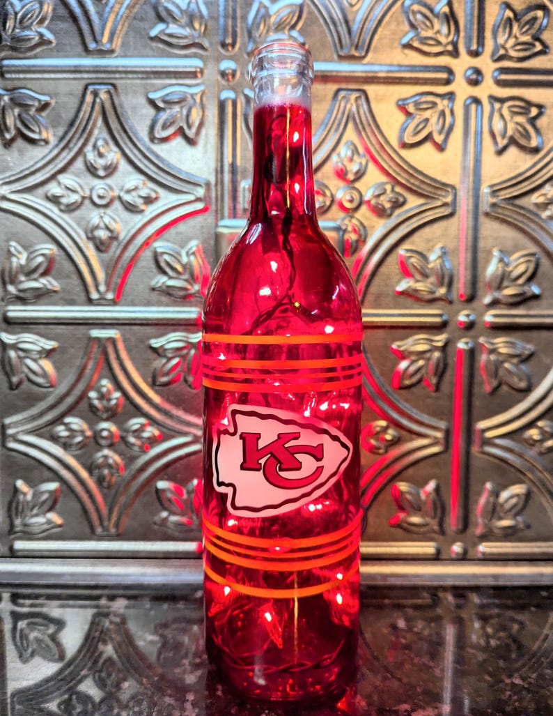 May include: A red glass wine bottle with a white and red Kansas City Chiefs logo. The bottle has orange stripes around it and is lit from inside with white lights.