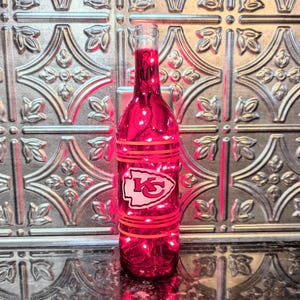 May include: A red glass wine bottle with a white Kansas City Chiefs logo and red stripes. The bottle is lit from inside with white lights.