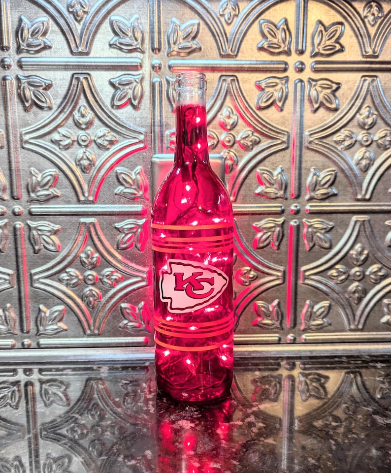 May include: A red glass wine bottle with a white Kansas City Chiefs logo and red stripes. The bottle is lit from inside with white lights.