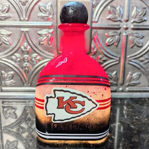 May include: A red glass bottle with a black top and a white and red Kansas City Chiefs logo. The bottle is decorated with black and white stripes and the word "PATRON" is printed on the bottom.