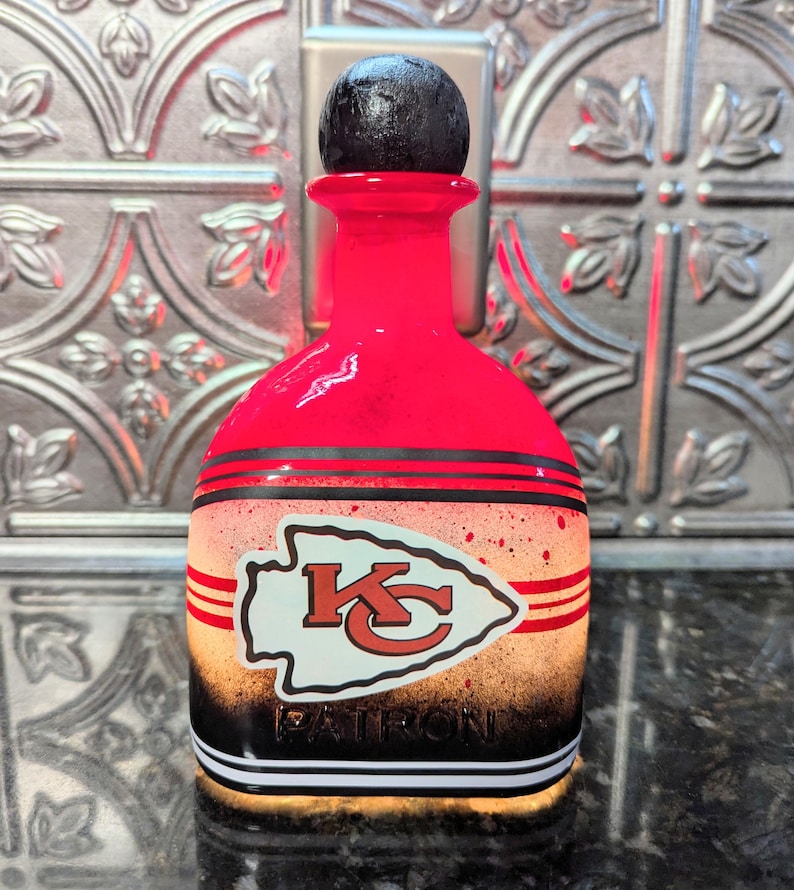 May include: A red glass bottle with a black top and a white and red Kansas City Chiefs logo. The bottle is decorated with black and white stripes and the word "PATRON" is printed on the bottom.