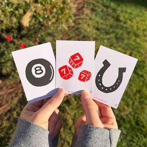 May include: Three rectangular white cards featuring black and red illustrations. The left card shows a black eight ball, the center card displays three red dice with the number seven, and the right card depicts a black horseshoe with small dots.