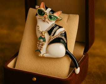 Op de afbeelding: Decoratieve kattenbroche in een fluwelen doos. De broche toont een witte kat met zwarte strepen, een groene zonnebril en een gouden ketting met een groene hanger. De kat is versierd met strass-steentjes.