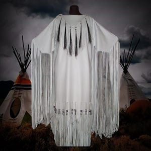 May include: A white dress with long fringe sleeves and hem, adorned with black tassels and decorative beading. The dress has a V-neckline and a flowing silhouette. The background features teepees and a cloudy sky, suggesting a Native American theme.