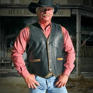 Könnte beinhalten: Eine schwarze Lederweste mit dekorativen Nähten und Knopfleiste. Die Weste wird über einem rosa Langarmhemd getragen und mit Jeans kombiniert. Ein schwarzer Cowboyhut vervollständigt das Outfit im Western-Stil.