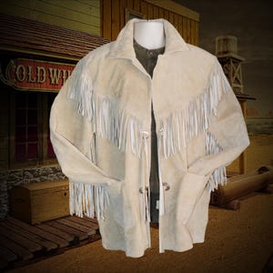 Könnte beinhalten: Cremefarbene Wildlederjacke mit weißen Fransen. Die Jacke im Western-Stil hat einen Kragen, zwei silberfarbene Knöpfe und Fransen an Ärmeln und Brust. Ein klassischer Western-Look.