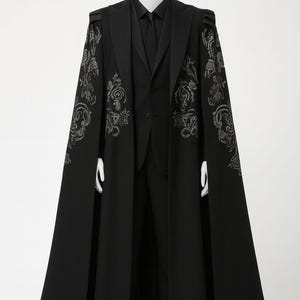 Puede incluir: Un traje negro con una capa larga. La capa presenta bordados florales plateados en los hombros y las mangas. El traje incluye una chaqueta negra, chaleco, camisa, corbata y pantal&oacute;n. El conjunto se exhibe en un maniqu&iacute;.