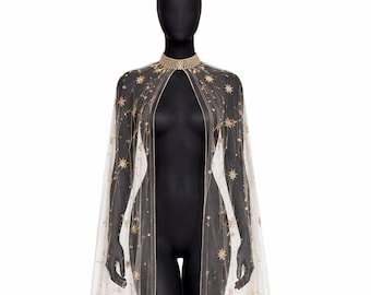 Peut inclure: Une cape transparente ivoire ornée d'étoiles et de points dorés. Elle présente un col haut orné et une longue conception fluide. La cape est présentée sur un mannequin noir sur fond blanc.