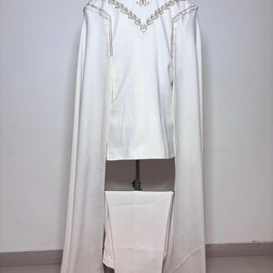 Puede incluir: Un traje blanco con una larga capa, exhibido en un maniqu&iacute;. La chaqueta presenta bordados dorados en la espalda y a lo largo de las costuras. Los pantalones son de corte recto y combinan con el color de la chaqueta. La capa se extiende desde los hombros.