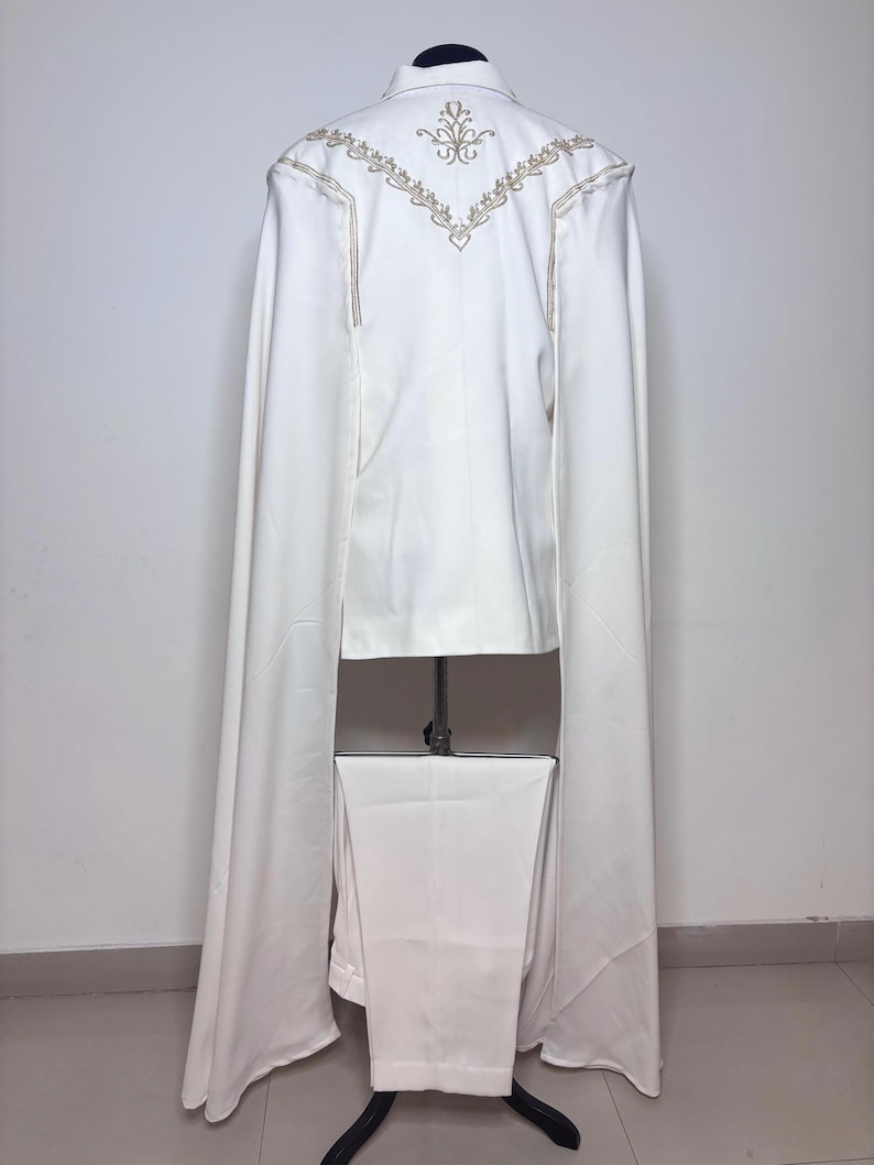 Puede incluir: Un traje blanco con una larga capa, exhibido en un maniqu&iacute;. La chaqueta presenta bordados dorados en la espalda y a lo largo de las costuras. Los pantalones son de corte recto y combinan con el color de la chaqueta. La capa se extiende desde los hombros.
