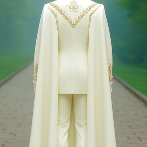 Puede incluir: Traje marfil con capa, exhibido en un maniqu&iacute;. El traje presenta bordados dorados en la espalda y las mangas. La capa fluye por la espalda, creando un aspecto formal y elegante. El fondo es una escena exterior borrosa.