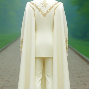 Puede incluir: Traje marfil con capa, exhibido en un maniqu&iacute;. El traje presenta bordados dorados en la espalda y las mangas. La capa fluye por la espalda, creando un aspecto formal y elegante. El fondo es una escena exterior borrosa.