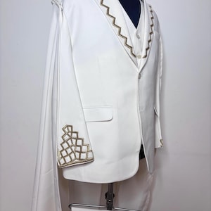 Puede incluir: Un traje blanco con bordados geom&eacute;tricos dorados en los pu&ntilde;os y el cuello. El traje incluye una blazer con capa, un chaleco y pantalones a juego. El traje se exhibe en un maniqu&iacute;.