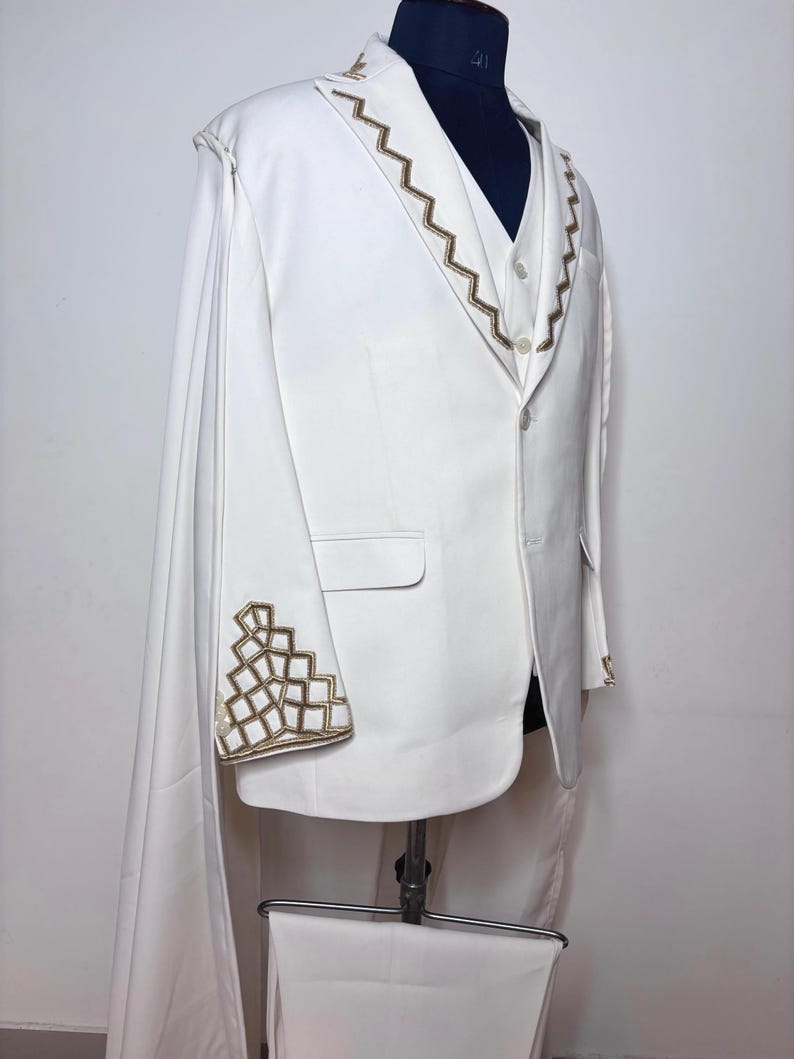 Puede incluir: Un traje blanco con bordados geom&eacute;tricos dorados en los pu&ntilde;os y el cuello. El traje incluye una blazer con capa, un chaleco y pantalones a juego. El traje se exhibe en un maniqu&iacute;.