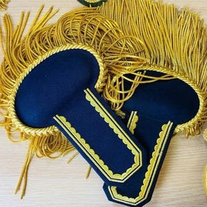 May include: Two navy blue epaulettes with gold trim and a gold rope border. The epaulettes are surrounded by gold fringe. The design is likely for a military uniform or formal wear.