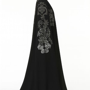 Puede incluir: Una larga capa negra con bordados plateados ornamentados en los hombros y las mangas. La capa se exhibe en un maniqu&iacute;, mostrando su dise&ntilde;o fluido y sus adornos detallados. La prenda parece estar hecha de una tela suave y elegante.