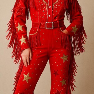 May include: Red suede jumpsuit with gold star and fringe detailing. The western-style outfit features a collared shirt with a button-up front, a matching belt, and flared pants. Gold embellishments and fringe adorn the sleeves and pant legs.