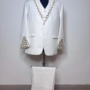 Puede incluir: Un traje de tres piezas blanco exhibido en un maniqu&iacute;. El traje presenta bordados geom&eacute;tricos dorados en los pu&ntilde;os y solapas. La chaqueta tiene un cierre de un bot&oacute;n, y los pantalones a juego est&aacute;n cuidadosamente doblados debajo.