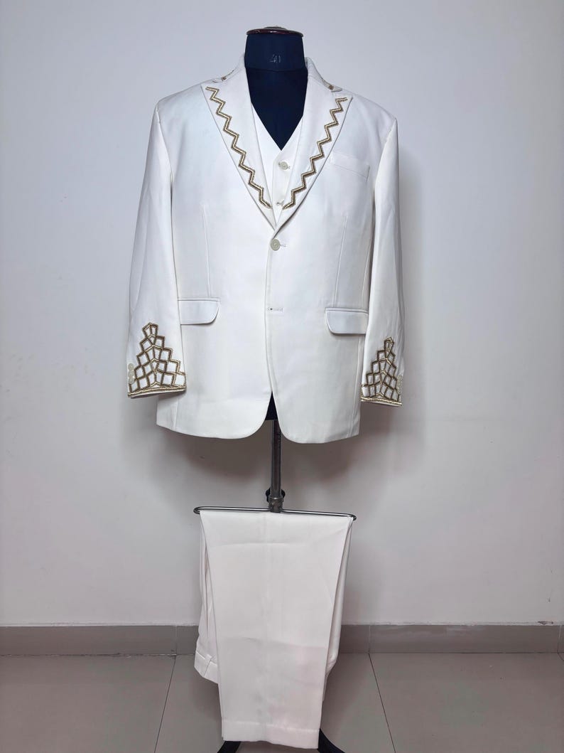 Puede incluir: Un traje de tres piezas blanco exhibido en un maniqu&iacute;. El traje presenta bordados geom&eacute;tricos dorados en los pu&ntilde;os y solapas. La chaqueta tiene un cierre de un bot&oacute;n, y los pantalones a juego est&aacute;n cuidadosamente doblados debajo.