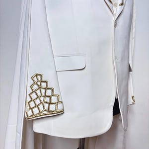 Puede incluir: Chaqueta de traje blanca con bordados geom&eacute;tricos dorados en la manga y el cuello. La chaqueta tiene un dise&ntilde;o tipo capa y se exhibe en un maniqu&iacute;. El traje es una prenda formal.