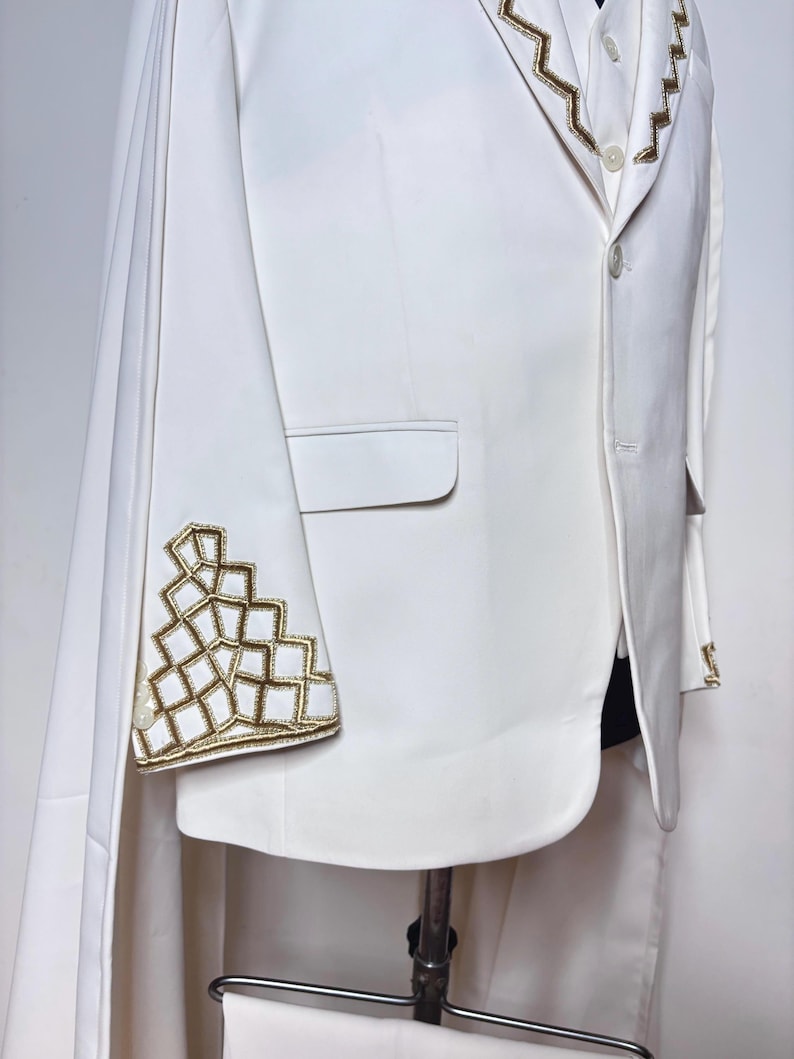 Puede incluir: Chaqueta de traje blanca con bordados geom&eacute;tricos dorados en la manga y el cuello. La chaqueta tiene un dise&ntilde;o tipo capa y se exhibe en un maniqu&iacute;. El traje es una prenda formal.