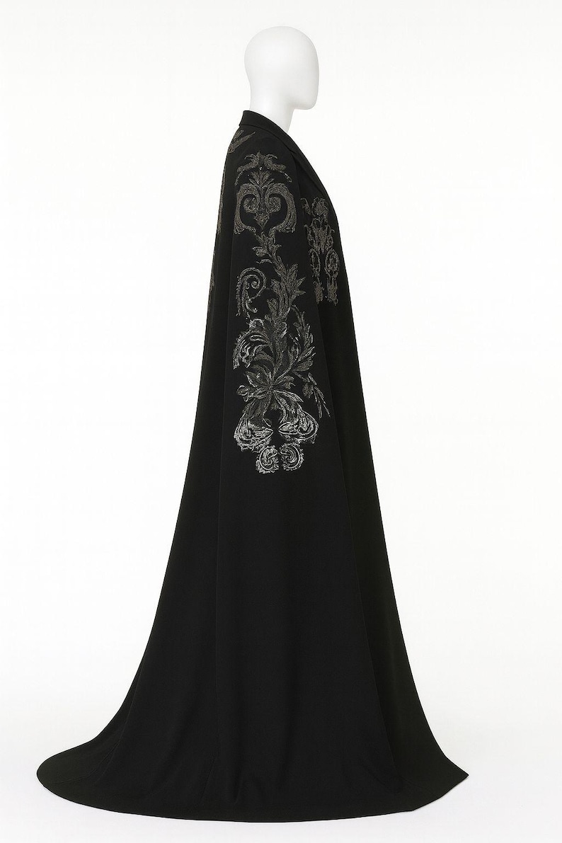 Puede incluir: Una larga capa negra con bordados plateados ornamentados en los hombros y las mangas. La capa se exhibe en un maniqu&iacute;, mostrando su dise&ntilde;o fluido y sus adornos detallados. La prenda parece estar hecha de una tela suave y elegante.