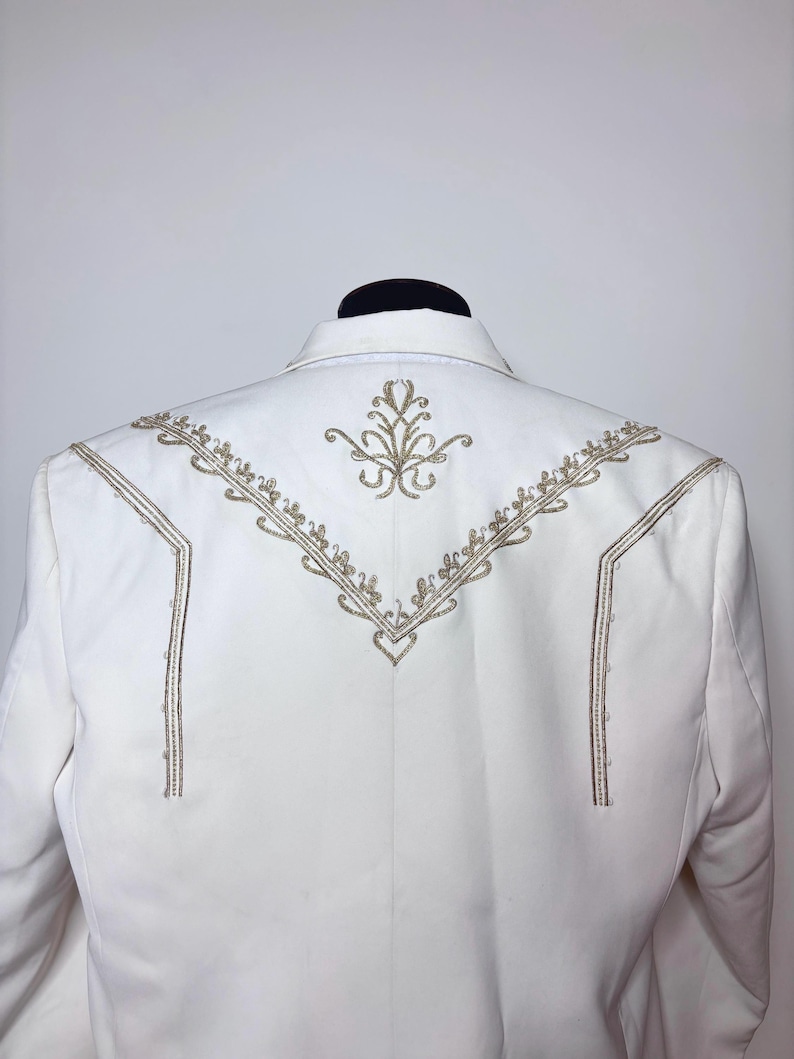 Puede incluir: Un blazer blanco con intrincados bordados dorados en la espalda. El dise&ntilde;o presenta un motivo floral central y l&iacute;neas decorativas. El blazer se muestra sobre una cabeza de maniqu&iacute; negra. Medidas en cent&iacute;metros.
