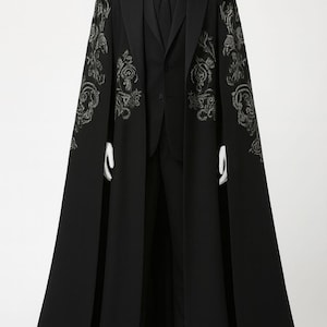 Puede incluir: Un traje negro con una capa larga. La capa presenta bordados florales plateados ornamentados en los hombros y las mangas. El traje incluye una chaqueta negra, chaleco, camisa, corbata y pantal&oacute;n. El conjunto se exhibe en un maniqu&iacute;.